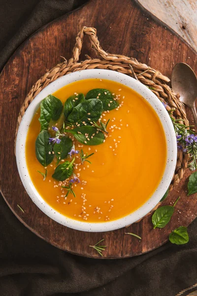 Canlı bir turuncu sebze püresi yeşil yapraklar ve tohumlarla süslenmiş benekli bir kasede servis edilir. Kır manzarası sıcak ahşap dokular ve doğal güneş ışığı içerir..