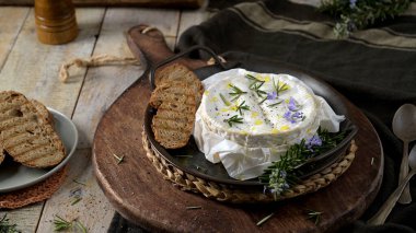 Sıcak, erimiş bir Camembert çarkı taze biberiye ve zeytinyağıyla süslenir. Sahne, kabuklu ekmek ve kırsal masanın dokusunu vurgulayan yumuşak, doğal ışıklandırmayı içeriyor..