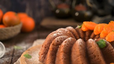 Kırsal bir turuncu demet kek, canlı mandalina segmentleri ve nane ile süslenmiş ahşap bir tahtanın üzerinde oturur. Sıcak, karamsar ışık yumuşak kırıntı dokusunu ve şekerin ince tozunu vurgular..