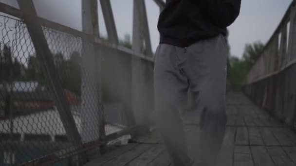 Aktiver junger Mann tanzt auf der Brücke, verschwommene Bäume und Himmel hinter ihm. — Stockvideo