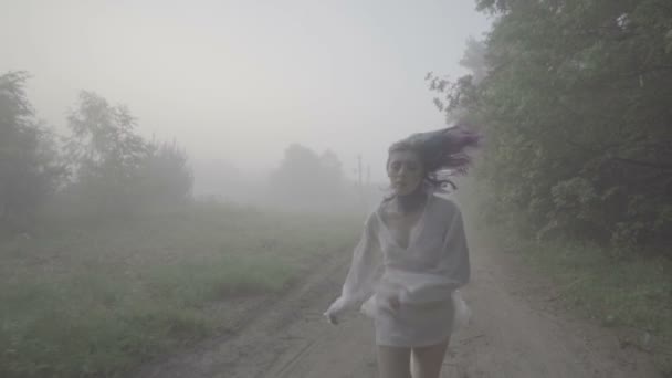 Vista frontal de la hermosa mujer de pelo morado y camisa blanca corriendo en el bosque - escena de suspenso. Video de belleza sensual entre árboles en cámara lenta, niebla y fondo oscuro del bosque . — Vídeo de stock