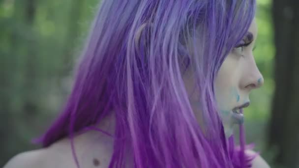 Primer plano de una mujer guapa con el pelo morado manchando su cara con pintura azul y mirando a la cámara. Aislado en el bosque, vídeo 4k . — Vídeos de Stock