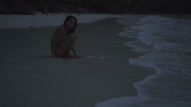 Mujer feliz hermosa asiática en bikini de ganchillo sentado en la playa por la sede. Video de la chica sonriente sexy sentada en la playa en cámara lenta . — Vídeos de Stock