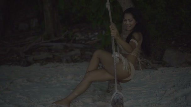 Mujer feliz hermosa asiática en bikini de ganchillo en la playa. 4k video de la chica sonriente sexy sentado en el columpio en la playa - vista lateral en cámara lenta . — Vídeos de Stock