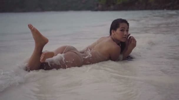 アジアの美しい女性かぎ針編みビキニを着てビーチで横になっています。水しぶきとスローモーションで動いて肉感的で砂の上に横たわるセクシーな女の子のビデオ. — ストック動画