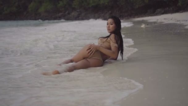 Mujer hermosa asiática en bikini de ganchillo acostado en la playa. Video de chica sexy tumbada en la arena con agua salpicada y tocando sensualmente - vista trasera en cámara lenta . — Vídeos de Stock