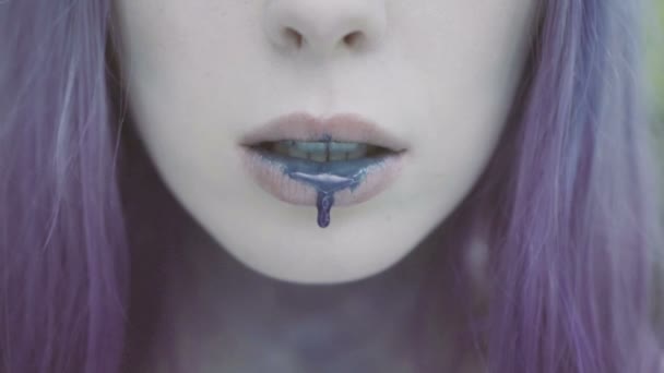 Close up of pretty woman face in purple hair with blue paint on her lips and drop falling down. Stationary shot of blue drop dripping from bottom lip. — Stock Video