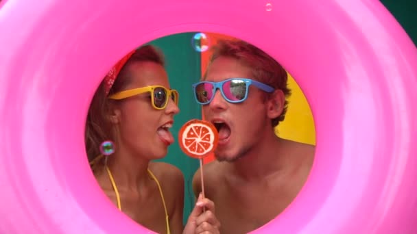 Funny young couple eating soap bubbles and licking orange lollipop isolated in the middle of pink inflatable ring and colorful wall in slow motion. — Stok video