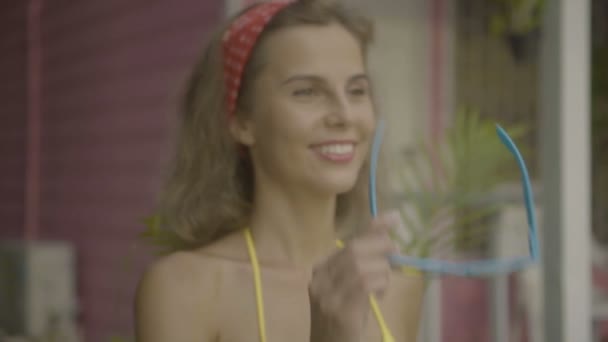 Pretty young happy woman wearing bikini taking off and putting on other sunglasses isolated on the background of pink house. — Stock Video