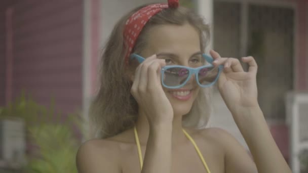 Mujer feliz bastante joven usando bikini poniéndose gafas de sol aisladas en el fondo de la casa rosa . — Vídeos de Stock