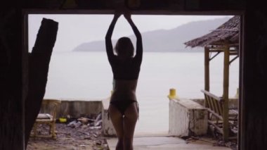 Arkadan görünüşü deniz arka plan - ağır çekimde video üzerinde terk edilmiş binada Teras penceresinde bir siyah şık mayo ayakta giymiş seksi sarışın kadın.