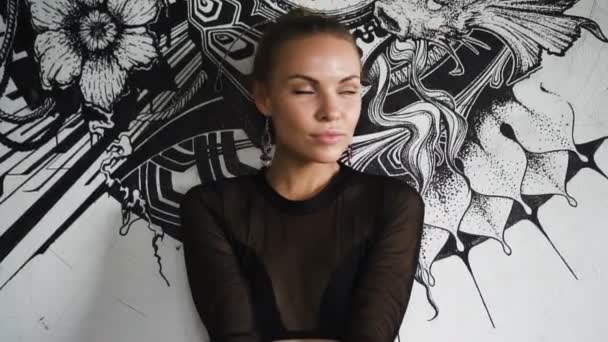 Sexig blond kvinna klädd i en svart elegant baddräkt och örhängen poserar i övergiven byggnad över väggen med svart och vit målning bakgrund. — Stockvideo