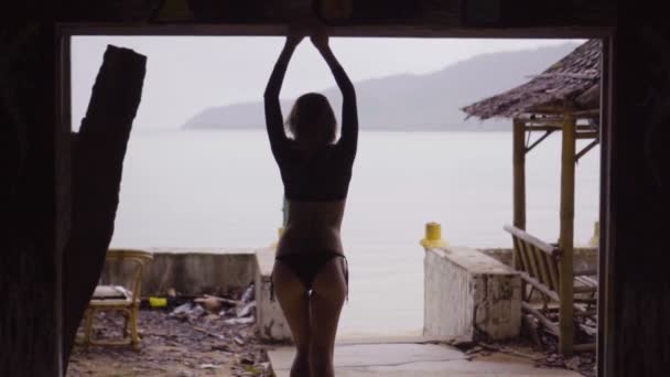 Bakifrån av sexig blond kvinna som bär en svart elegant baddräkt stående i övergiven byggnad terrass fönster över havet bakgrund - video i slow motion. — Stockvideo