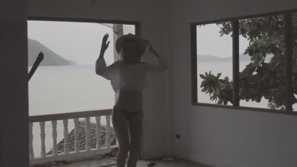 Ziemlich natürlich aussehende Frau an einem Sommertag im Boho-Stil mit weißem Hemd, Bikini und Hut tanzt in einem verlassenen Gebäude vor wunderschönem Meereslandschaftshintergrund. — Stockvideo
