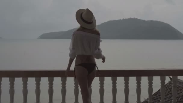 Back view of pretty naturally looking woman on summer day in boho style fashion wearing white shirt, bikini and hat posing and smiling on a terrace over beautiful sea landscape background. — Stock Video