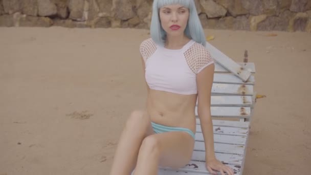 性感的美丽女人在现代未来派风格的视频，在损坏的木制蓝色日光浴床上摆姿势。穿着比基尼和蓝色假发在海滩上的女人的创意外观. — 图库视频影像