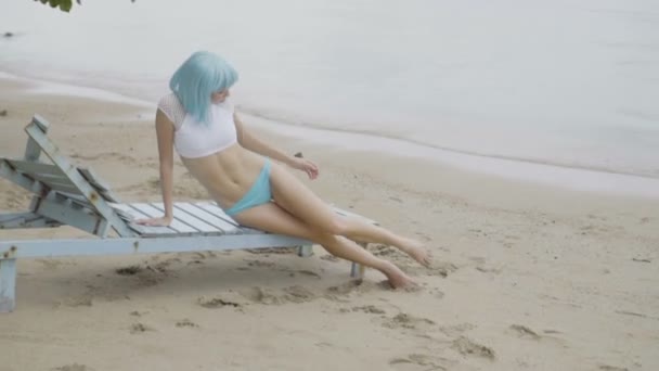 손상된 나무 파란색 일광욕용 침대에 포즈를 취하는 현대 미래 지향적 인 스타일의 섹시한 아름다운 여성의 비디오. 비키니와 파란색 가발을 입고 해변에서 여자의 창조적 인 모습. — 비디오