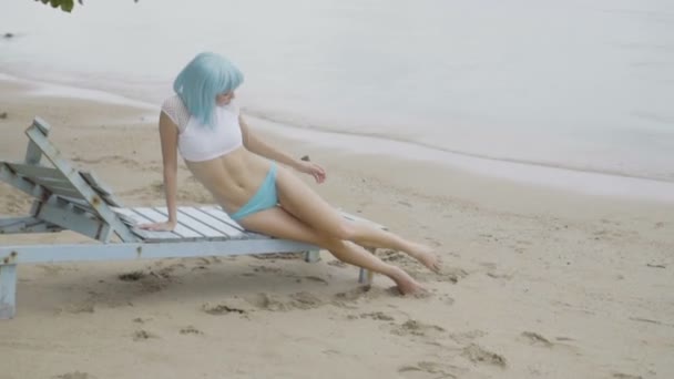 Video av sexig vacker kvinna i modern futuristisk stil poserar på den skadade trä Blå solbädd. Kreativt utseende av kvinna på stranden klädd i bikini och blå peruk. — Stockvideo