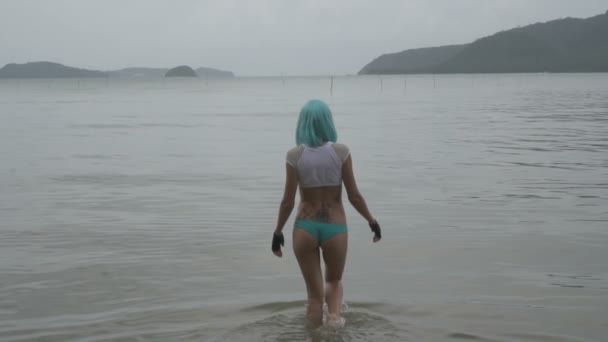 Video de sexy hermosa mujer en estilo futurista moderno caminando en el mar. Aspecto creativo de la mujer tatuada que usa bikini, peluca azul, guantes sin dedos de cuero negro en el agua - vista posterior en cámara lenta . — Vídeo de stock