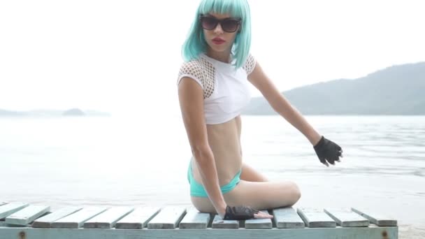 Vidéo de belle femme sexy dans un style futuriste moderne posant sur le transat en bois bleu sur fond de mer. Look créatif de femme sur la plage portant un bikini, une perruque bleue, des gants sans doigts en cuir noir et des lunettes de soleil - vidéo au ralenti . — Video