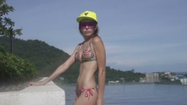 Bikini, sarı hat ve sonsuzluk çatıdaki yüzme havuzunda güneşli bir günde mavi gökyüzü ve yeşil ağaçlar manzara üzerinde poz mor güneş gözlüğü giyen genç ve güzel kadın.