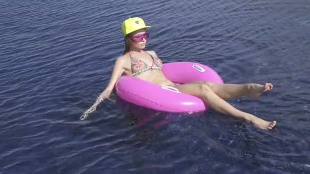 Pretty young woman wearing bikini, yellow hat and purple sunglasses sitting on a pink inflatable ring in swimming pool on a sunny day. — Stock Video
