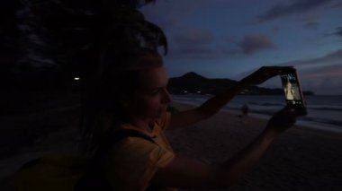 Plajda gün batımı sırasında cep telefonu ile selfie fotoğraf çeken kadının silueti.