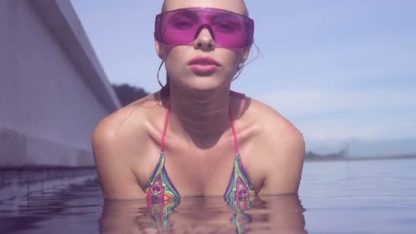 Face closeup de sexy jovem mulher vestindo óculos de sol roxos olhando para a câmera na piscina infinito telhado em um dia ensolarado sobre céu azul e verde árvores paisagem . — Vídeo de Stock