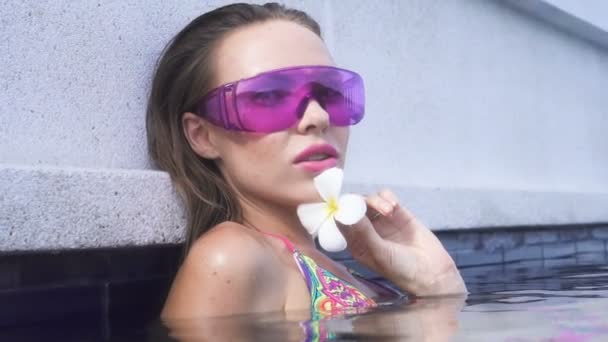 Primeros planos de la cara de la joven sexy que usa bikini y gafas de sol púrpura con flor blanca mirando a la cámara en la piscina infinita de la azotea en un día soleado . — Vídeo de stock