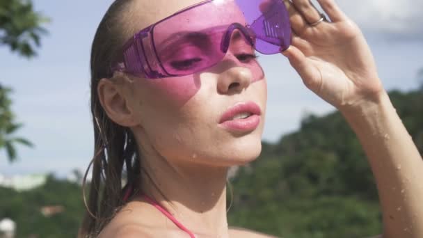 Face closeup de sexy jovem mulher vestindo óculos de sol roxos na piscina no telhado em um dia ensolarado sobre céu azul e verde árvores paisagem . — Vídeo de Stock