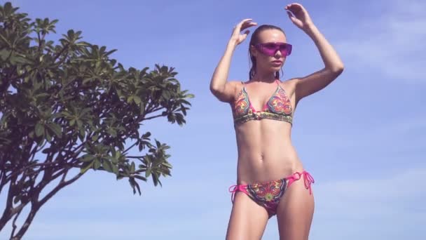 Sexy jonge vrouw dragen van een bikini en paars zonnebril poseren bij zwembad op het dak op een zonnige dag over blauwe hemel landschap. — Stockvideo