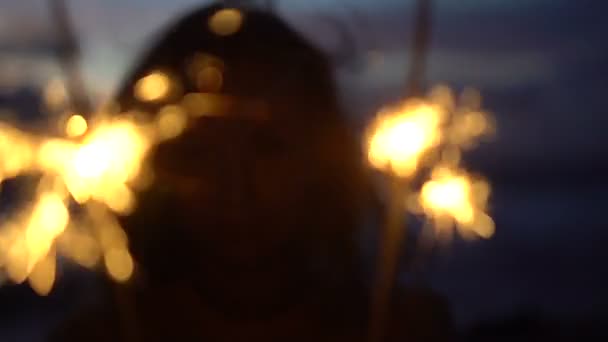 Primer plano de mujer feliz con fuegos artificiales chispeantes en la playa al atardecer . — Vídeos de Stock