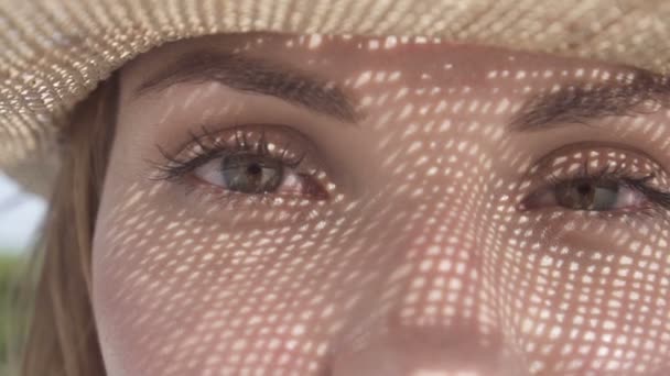 Retrato de una hermosa mujer joven con sombrero de paja en un día soleado mirando a la cámara sobre el cielo azul y el paisaje de árboles verdes. Ojos primer plano de chica bonita con sombrero de paja patrón sombra en su cara . — Vídeos de Stock