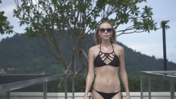 Sexy blonde glamour woman wearing black swimwear and sunglasses walking on a rooftop in a sunny summer day with beautiful sky and green mountain landscape background - video in slow motion. — Stock Video