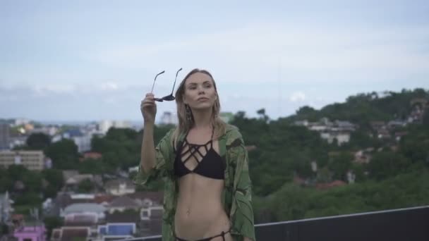 Sexy blonde Glamour-Frau trägt schwarze Badebekleidung und florales langes Hemd, als sie an einem sonnigen Sommertag mit schönem Himmel, grünem Berg- und Stadtlandschaftshintergrund auf einem Dach spaziert - Video in Zeitlupe. — Stockvideo