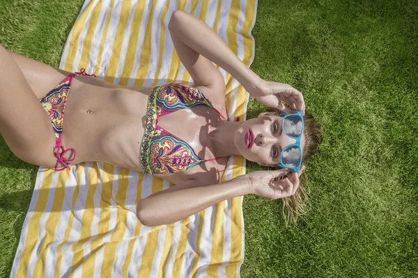 Felülnézet szexi fiatal nő visel bikini és a kék napszemüveg feküdt csíkos strand törülközőt, mint a zöld fű háttér — Stock Fotó