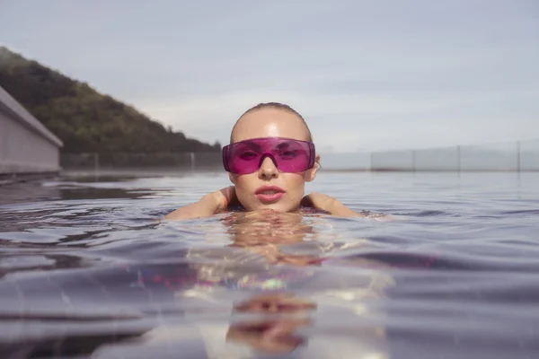 Face closeup of sexy young woman wearing purple sunglasses looking at camera in infinity rooftop swimming pool on a sunny day over blue sky and green trees landscape — Stock Photo, Image