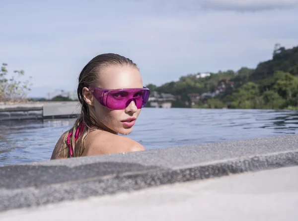 Primer plano de la cara de la joven sexy con gafas de sol púrpura mirando a la cámara en la piscina infinita en la azotea en un día soleado sobre el cielo azul y el paisaje de árboles verdes — Foto de Stock