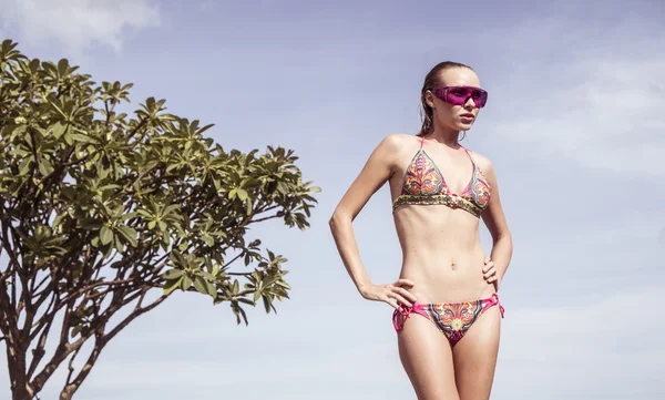 Szexi fiatal nő pózol a tetőtéri medence napos táj kék ég alatt, a bikini és a lila napszemüveget visel. — Stock Fotó