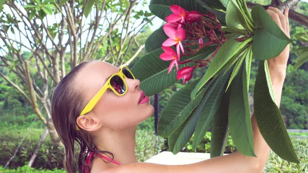 Primeros planos de una joven sexy que usa bikini y gafas de sol amarillas con cabello húmedo oliendo a flor de árbol en la azotea en un día soleado — Foto de Stock