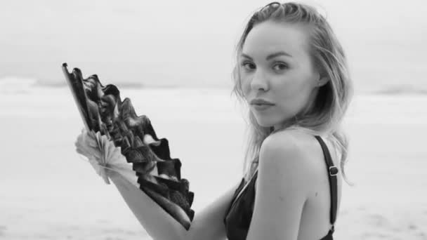 Piękna Blondynka Glamour kobieta ubrana w czarne stroje kąpielowe pozowanie na plaży w słoneczny letni dzień z chińskim składanym fanem dłoni nad morzem i tłem nieba-czarno-białe wideo w zwolnionym tempie — Wideo stockowe