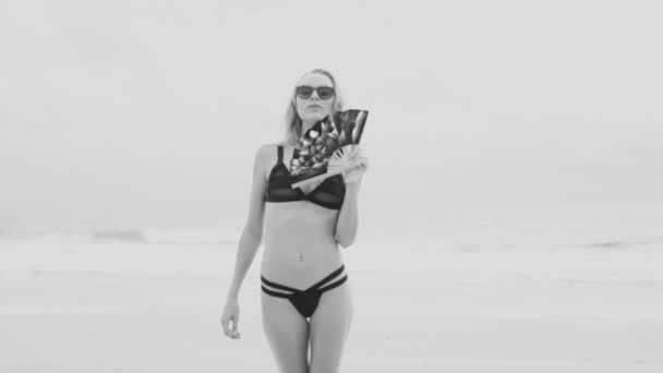 Hermosa mujer rubia glamurosa con traje de baño negro y gafas de sol posando en una playa en un día soleado de verano con ventilador de mano plegable chino sobre el fondo del mar y el cielo - vídeo en blanco y negro — Vídeo de stock