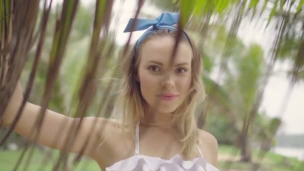 Güzel bir yaz günü yürüyüş ve palmiye ağaçları arasında gülümseyen beyaz bikini üst ve mavi kafa bandı yay giyen oldukça doğal görünümlü kadın Closeup - yavaş hareket video — Stok video