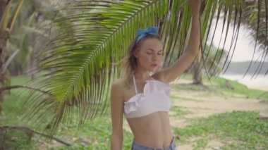 Güzel bir yaz günü palmiye ağaçları arasında yürüyüş beyaz bikini üst, kot şort ve mavi kafa bandı yay giyen Pretty doğal görünümlü kadın - yavaş hareket video