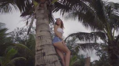 Beyaz bikini üst giyen oldukça doğal görünümlü kadın alt görünümü, kot şort ve mavi kafa bandı yay güzel bir yaz günü ayakta, bir palmiye ağacına yaslanmış - yavaş çekimvideo