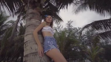 Beyaz bikini üst giyen oldukça doğal görünümlü kadın alt görünümü, kot şort ve mavi kafa bandı yay güzel bir yaz günü ayakta, bir palmiye ağacına yaslanmış - yavaş çekimvideo