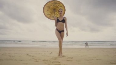 Çin şemsiyesi gülümseyen ve plajda güzel bir yaz gününde poz siyah mayo giyen güzel sarışın glamour kadın alt görünümü - yavaş hareket video