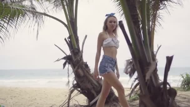 Vrij natuurlijk uitziende vrouw dragen witte Bikinitop, jeans shorts en blauwe hoofdband Bow op een mooie zomerdag wandelen en kijken naar de camera tussen palmbomen — Stockvideo