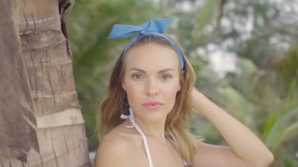 Красивая естественно выглядящая женщина в белом топе бикини и синей повязке на голове в прекрасный летний день улыбается, смотрит в камеру и стоит, опираясь на пальму - видео в замедленной съемке — стоковое видео
