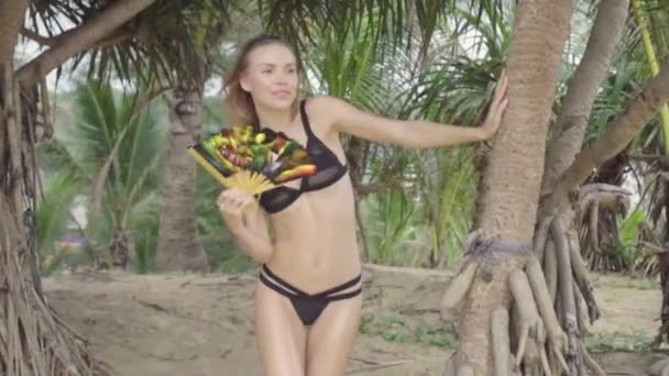 美丽的金发魅力女人穿着黑色泳装微笑，并摆出一个美丽的夏日与棕榈树之间的中国折叠手扇 - 视频在慢动作 — 图库视频影像